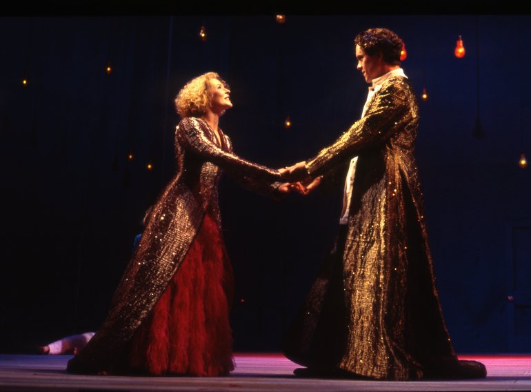 Two of the lovers hold both hands, but at arms' length. The fair-haired lady wears a red full-length dress and an embroidered gold cloak; the dark-haired man, who is a head taller, is also wearing  gold embroidered clothes.