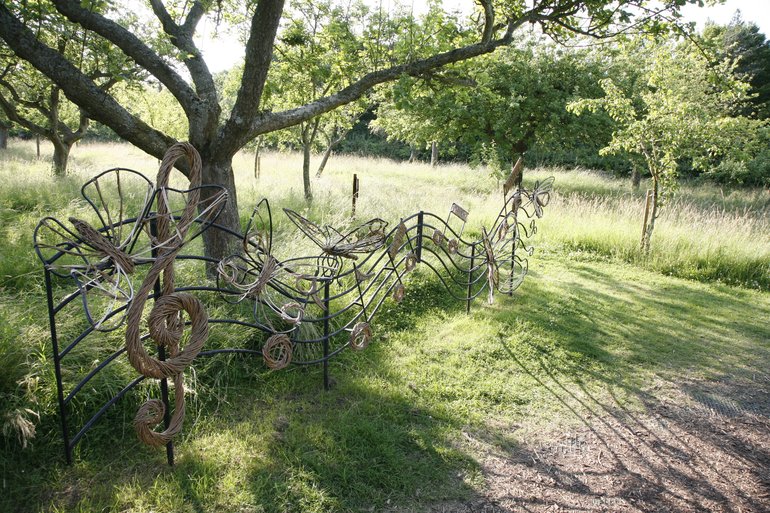 A metal musical stave runs in a wave away from the viewer. On it are metal butterflies and musical notes, with a treble clef at the beginning. Behind are the trees of an orchard standing in long grass, with a mown path beside the sculpture.