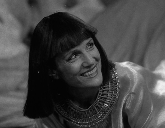 A smiling female with straight black hair with a straight fringe leans to her right and looks up to her left. She wears a rich decorative metal collar and a dress which reflects the light. (The picture is black and white.)