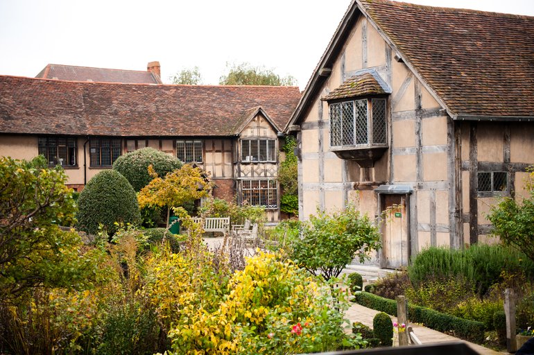 The rear wall of the Birthplace, with a jutting first-floor window, is on the right.. In front are shrubs and trimmed box trees, and a path leading to a group of benches, while in the rear is a row of timber-frame houses.