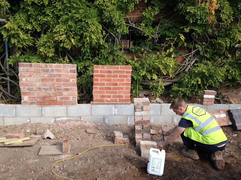 Brickwork options. On a low wall of white blocks are two samples - one of pale old-looking brick and the other of much redder modern brick. A workman crouching is preparing a third sample.