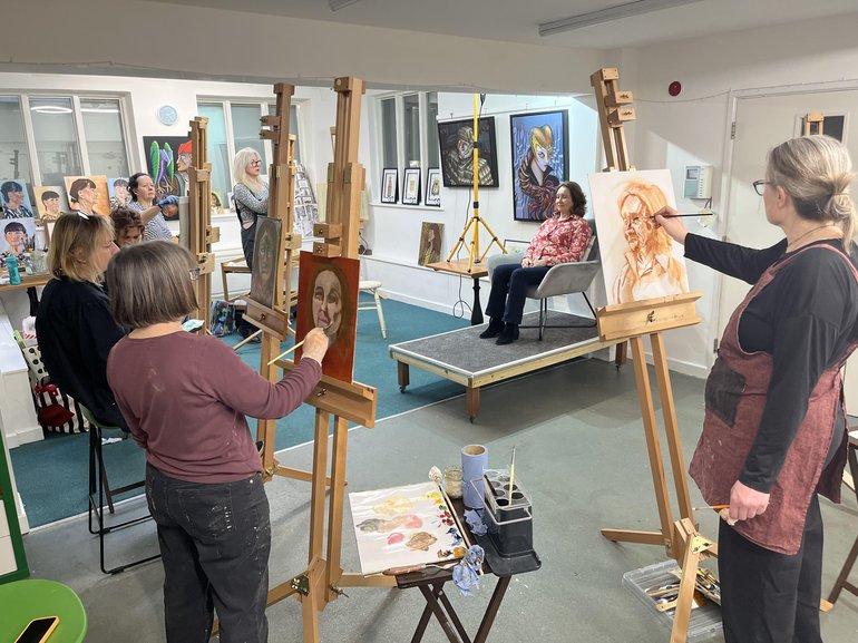 A group of artists stood or sat in front of easels with painting equipment around them, creating a portrait of a model who is sat in front of the group