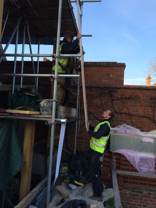 Scaffolding being put in place; two men are lifting boards to form an upper level.