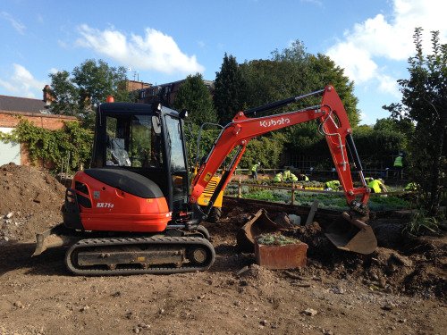 The image shows a large (stationary) red mechanical digger standing on bare earth. In the background people in yellow high-viz vests are working in the garden.