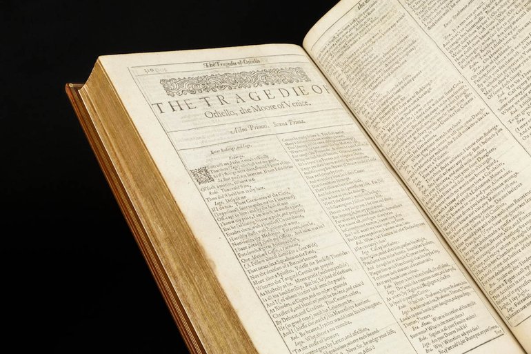The First Folio edition of Shakespeare's plays is open at the first page of Othello. The text is printed in small type in two columns, but the title spans the page, with a decorative bar above it. The volume is bound in brown leather.