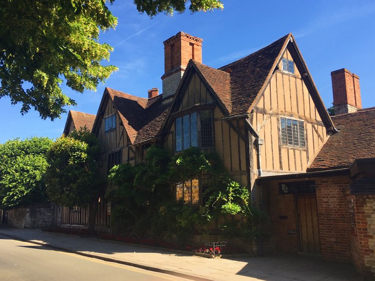 The front of the house with its three gables, large windows and tall red-brick chimneys. The house is in shade, but the nearest ground-floor window is lit. The sun is on the end wall, showing the large quantity of very straight timber beams.