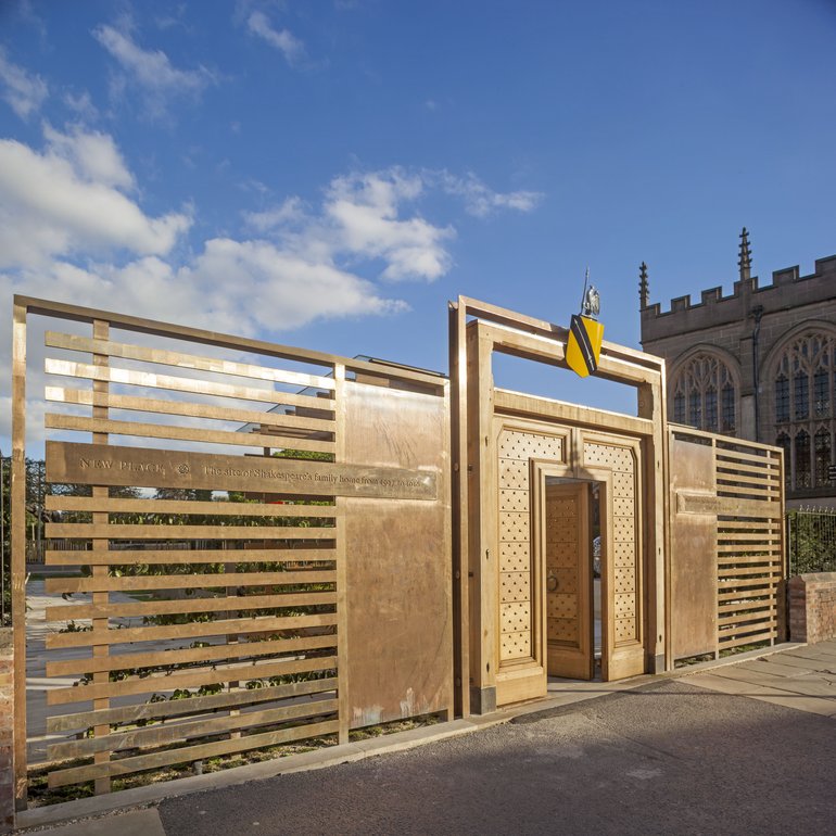 New Place gate house. A horizontally slatted fence recedes from left to right, with a door in the middle. This is surrounded by a patterned panel, and there is an outer frame extending above the height of the wall and surmounted by Shakespeare's arms.