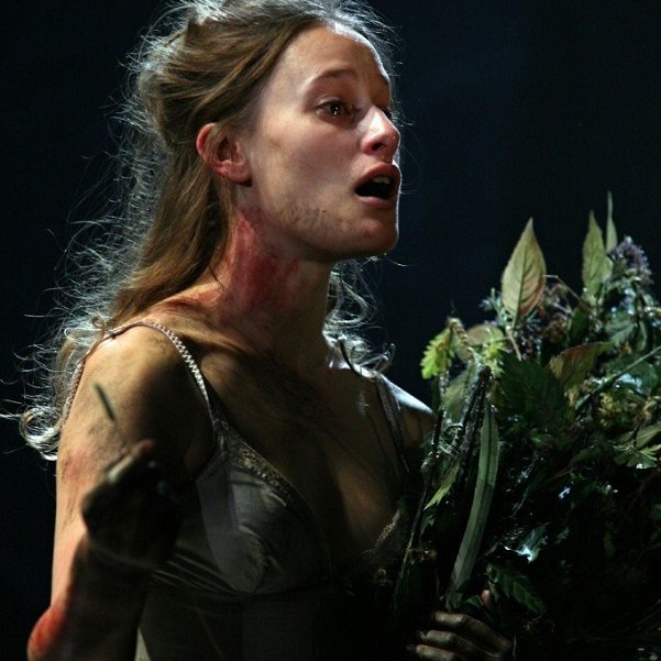 Against a dark background Ophelia, dramatically-lit, looks to the right with an open mouth. Her arms are bare, and her colouring looks highly bronzed. She is carrying a bunch of leaves.