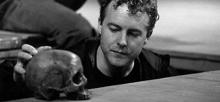 Hamlet, standing in a grave, contemplates the skull of Yorick resting on the edge of the grave; Hamlet's right hand is on the back of the skull.