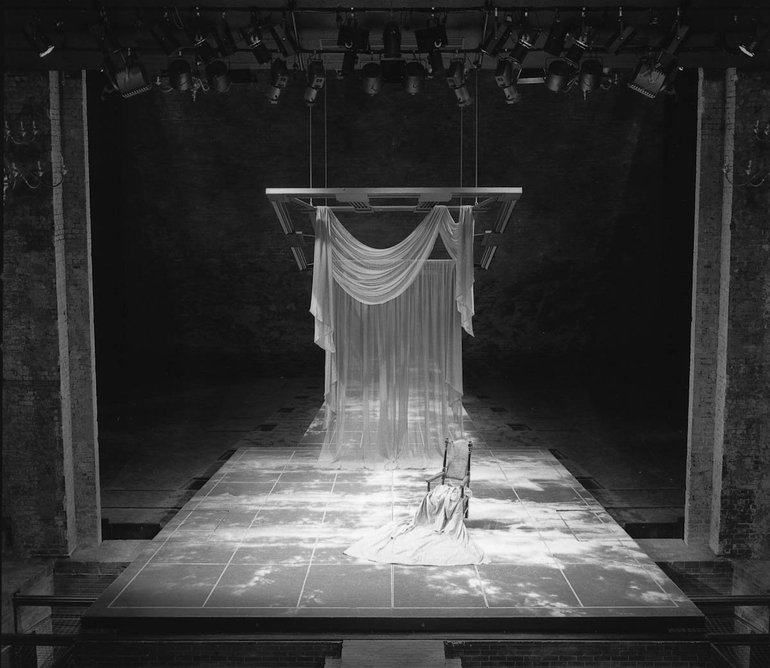 The set shows a largely empty stage. On a floor marked with squares, and which extends forward of the proscenium arch, is just a cloth-draped chair. Some way behind a semi-transparent curtain with a curved drape at the top hangs from a wooden frame.