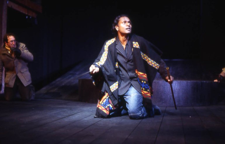 Hamlet, holding a sword and what may be either a dagger or Laertes' sword kneels looking up at his opponent (out of shot). He wears modern clothing, a suit jacket and jeans, but with African-style designs on the sleeves and one leg.