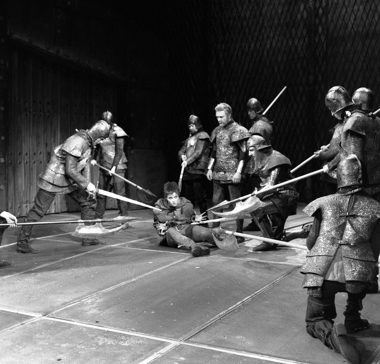 A number of men in armour and helmets point halberds and swords at a young man seated on the ground, while a bearded man without armour, wearing  a tunic with armorial  designs stares down at the young man.