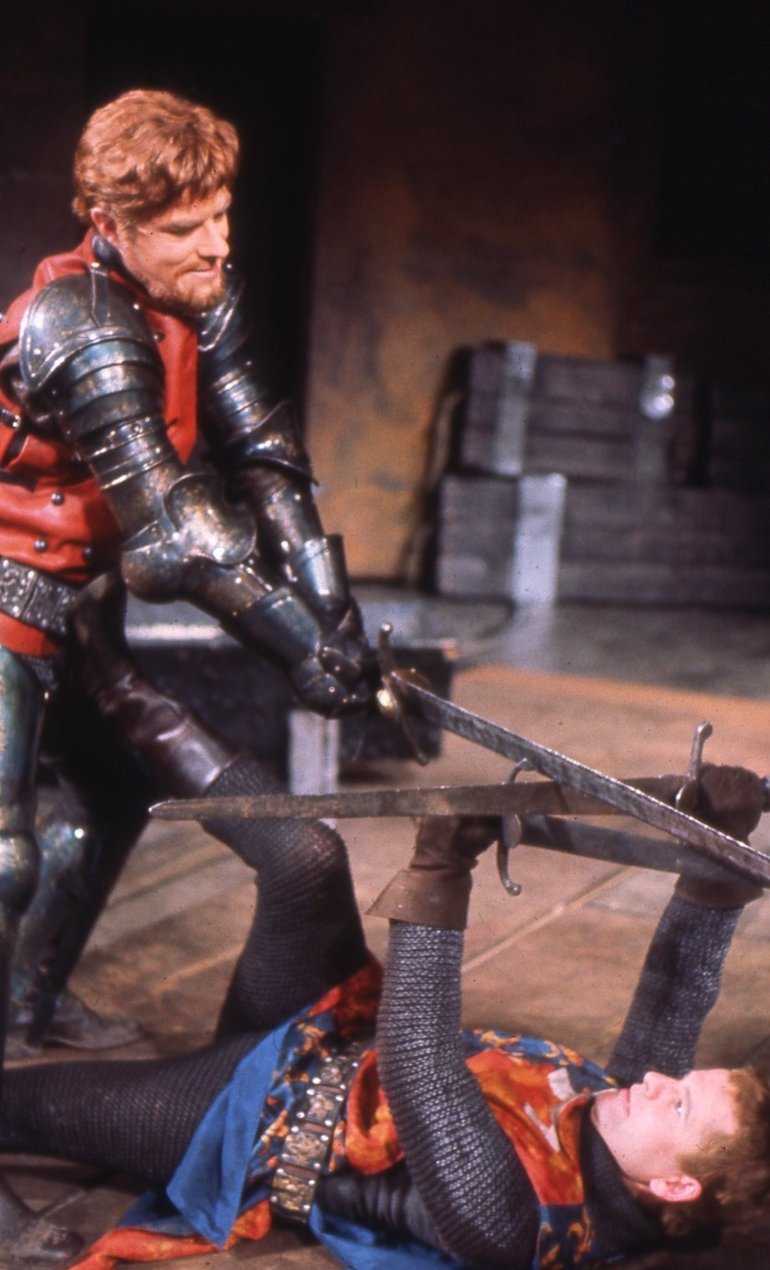 Douglas, with red hair and beard and wearing full armour and a red surcoat, Brings his sword down on the sword of a figure lying on the ground who is holding his sword up horizontally at arms length. He is wearing chain mail, with a royal surcoat .