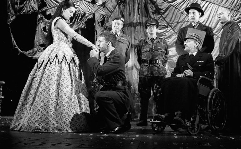 Henry V kisses the hand of Princess Katherine; she wears a full-length crinoline, he a military uniform. The French king, in a wheelchair, wears a French military uniform. In the background are two army officers and a diplomat in black hat and coat.