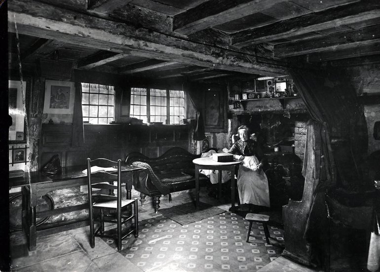 A black and white photo of Mary Baker sitting next to the Courting Settle in Anne Hathway's cottage. Mary sits at a round table, in a dark-coloured dress and white apron.
