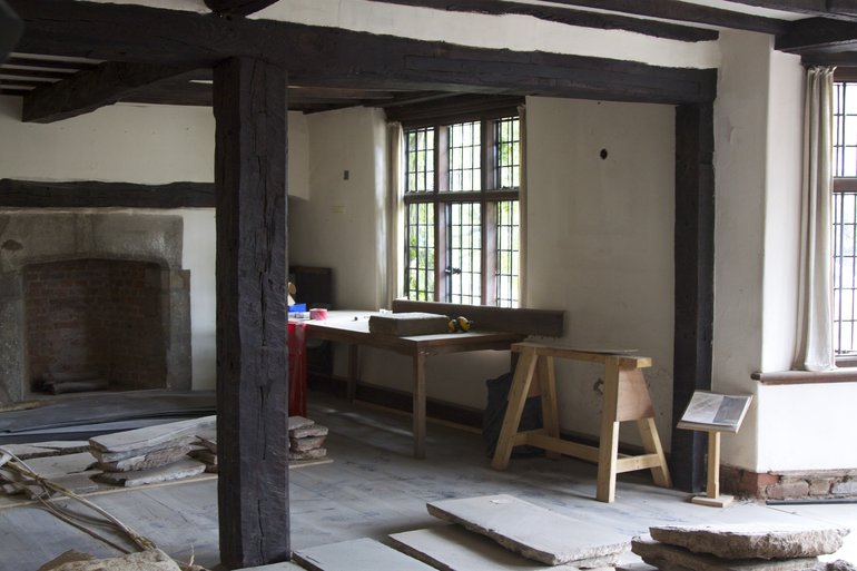 The image is of a room in a timber-framed house, with a vertical wooden beam in the foreground, part of a mid-room truss. There is a stone fireplace in the background,, and the windows are leaded. Building work is taking place.