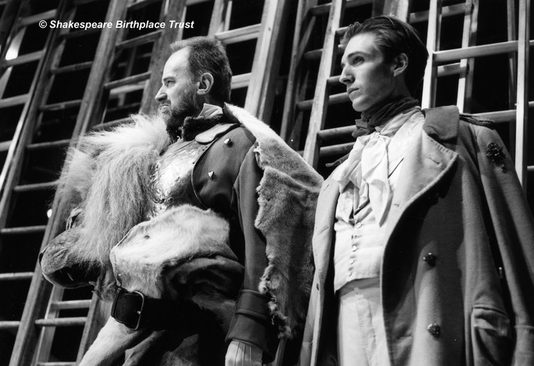 King John, RSC, 1988. Two men stand against a background of ladders. Both wear 19th century military uniforms; the older man has a furred cape over his shoulder. Both look grim as they stare to the left.