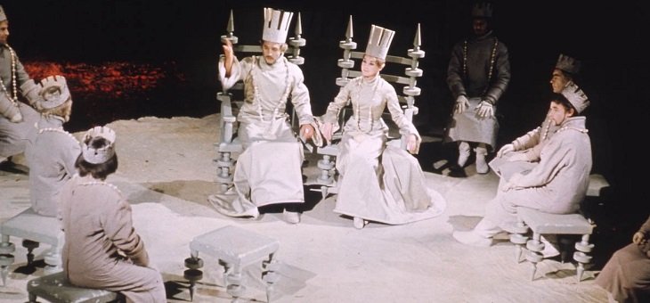 Macbeth Royal Shakespeare Company, 1967 (featuring funny hats). All characters are dressed in white. Macbeth and his lady sit on high-backed chairs facing us, other characters sit on stools around and facing them. All wear paper or cardboard crowns, Macbeth's and his lady's being taller than the rest.