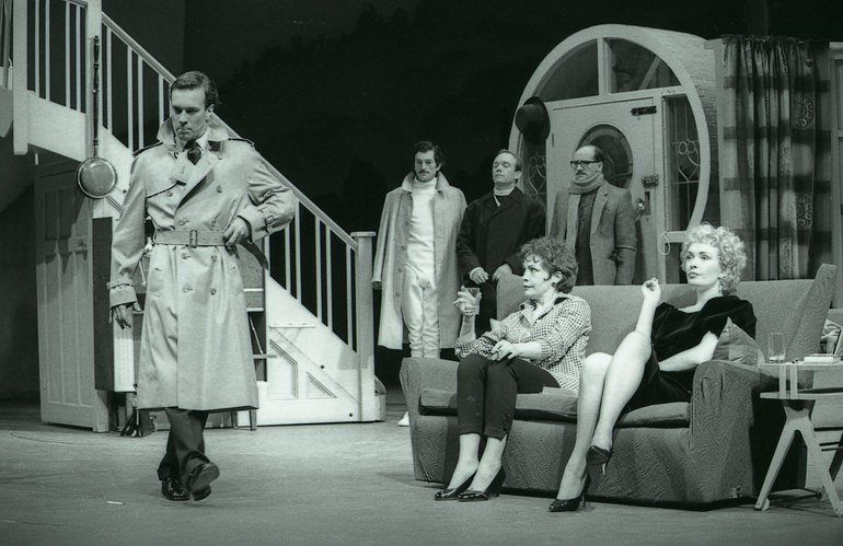 Merry Wives of Windsor, RSC, 1985 In modern dress, two ladies sit on a sofa while three men stand behind between bookshelves and a stair. All are looking disapprovingly at a man in a raincoat standing in front of the sofa, side-on to the ladies and looking downwards with a hurt expression.