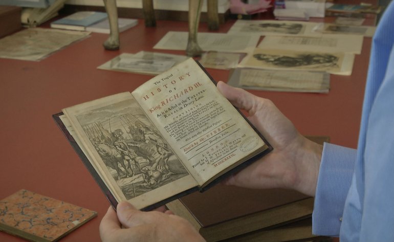 The picture shows a table covered with books and printed pages, and in the foreground the hands of a reader holding an open copy of an early edition of King Richard III, showing the title page and a frontispiece.