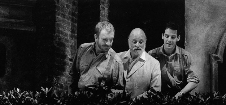 Three men, two young, one older, are looking at something below a hedge.
