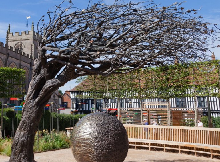 A dead tree on the left curves over a bronze globe with a pock-marked surface. In the background is a long timber-framed building on the right, and the Guild Chapel to the left. There is a clear blue sky.