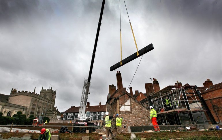 Against a dramatic backdrop of a sky with wild grey clouds and the chimneys , timber-framed houses and Guild Chapel surrounding New Place, a large plank of timber swings from a very tall, thin crane.