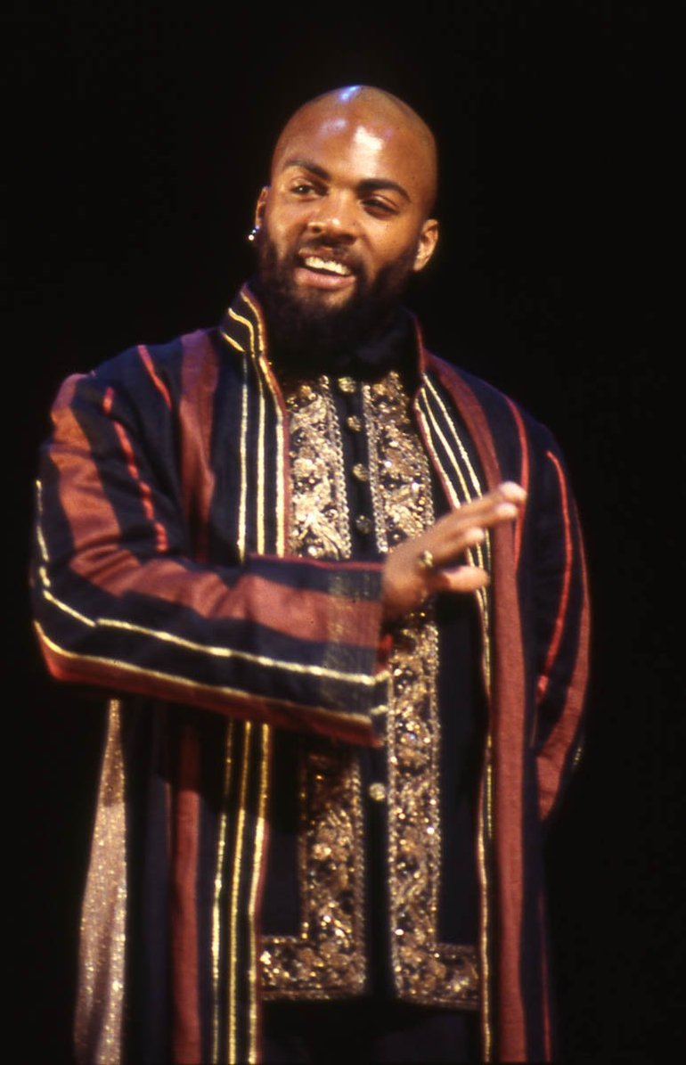 Othello Royal Shakespeare Company, 1999. A  black Othello wears an embroidered jacket and a brown gown with broad black and thin white stripes. He stand with his right arm across his body, gesturing rejection; his expression is disdainful.