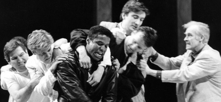 Two men in black with the appearance of front-row forwards, linked as in a scrum, carry another figure in white on their shoulders, with two other figures in white pushing the shoulder of one of the carriers. On the right an older man appears to be attempting to steady the load.