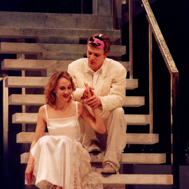 In modern dress, Juliet wears a while low-cut silk-looking dress and Romeo a white suit and a carnival mask which he has raised to his hairline. The sit on a set of wooden stairs, Juliet below and to the right of Romeo; her left hand is lifted and held in both of his. He looks seriously at her, while she looks modestly down, smiling.