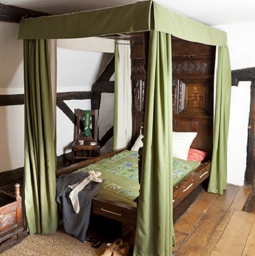 A four-poster bed with green hangings and a green coverlet on the bed. There is a solid oak cared headboard. on the bed are a pair of white knee-length socks and a green kirtle. In the background is a wooden chair, and everything stands on a wooden floor.
