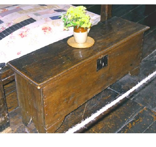 A plain oak chest with a lock; on it stands a stoneware jug on a circular rush mat. There are yellow flowers in the jug,  and part of  a bed can be seen in the background.