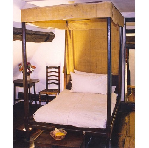 A four-poster bet with a canopy of yellow wool, extending about 20 centimetres down each of the posts. Yellow wool also covers the back. There are a chair and round  table with a vase of flowers in the background; in the foreground is a wooden chest .