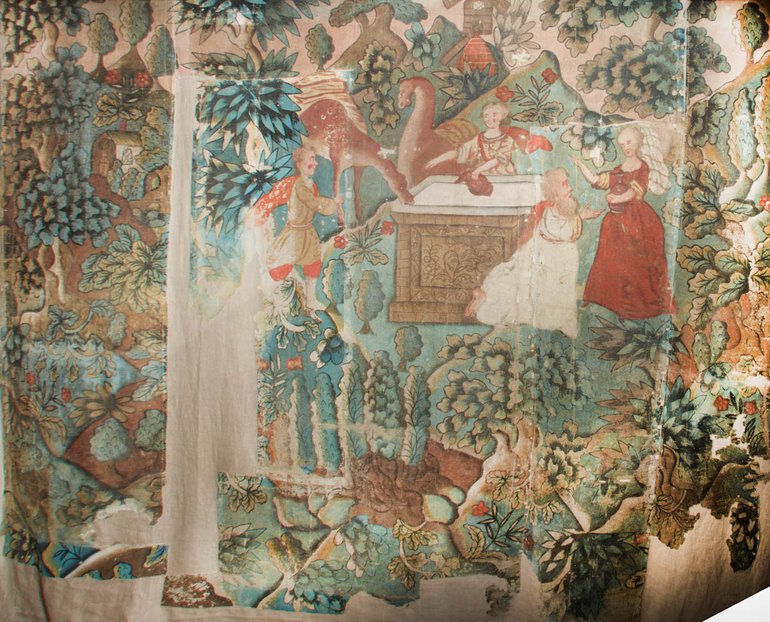 The highly-coloured wall hanging, imitating tapestry, shows a man holding a rabbit on a solid wooden table with a sunken stone top, and a woman in red greeting a child in white who runs to he. They are surrounded by plants and animals.