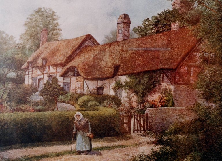Mary Baker stands on the pavement outside the front gate; she wears dark jacket over a white dress, a white bonnet tied under her chin, and she is resting on two sticks. Behind her the garden hedges are trimmed, and the cottage looks in good condition, with roses climbing the wall of the section nearest the road.