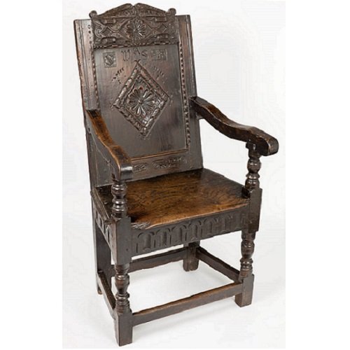 A wooden arm chair with carved panels between the legs supporting a leather-covered seat. The back has a highly-carved shaped top, and a carved border frames a panel with a flower-design in a diamond in its centre, and a small Shakespeare shield.