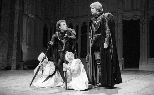 Richard, with a crutch and a withered arm, stands above two children in white. He is talking to a tall man wearing a cloak over a tunic.