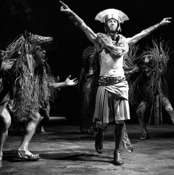 Three creatures with wolves' heads on top of their visible faces surround threateningly a bare-chested Caesar wearing a helmet with a transverse crest. An armoured and helmeted legionary is in the background. (2009 production)