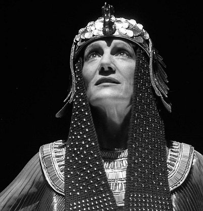 Harriet Walter as Cleopatra, wearing a cap of coloured discs, from which a black scarf with white spots hangs down either side of her face to her chest. She also wears a decorated metal collar over her shoulders.