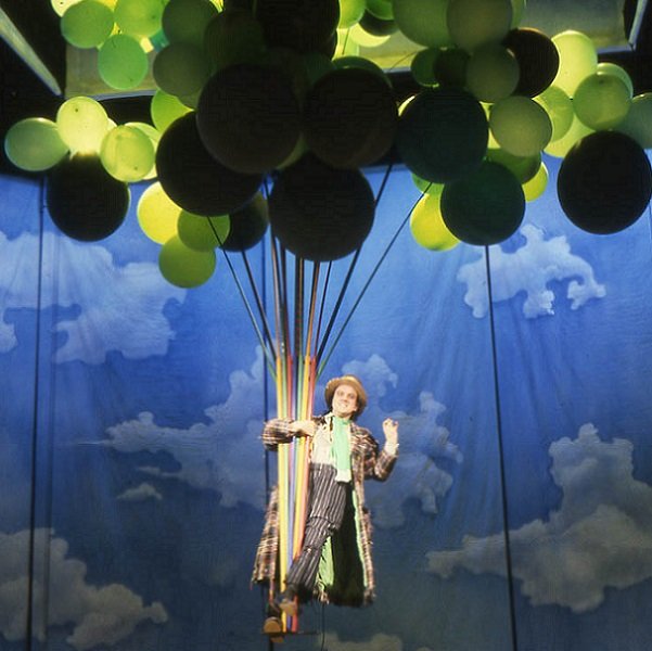 Against a background of darkish blue sky and white-ish clouds a man in a straw hat, checked trousers and checked coat flapping open is being held aloft by a large bunch of yellow and dark balloons. From a production of "As You Like It".
