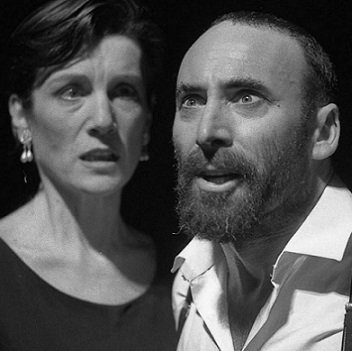 Macbeth and Lady Macbeth in modern dress. He is staring open-mouthed at something, while lady Macbeth looks sideways at him, with a worried face.