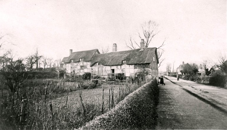 View of Anne Hathaway's Cottage from the road. At this time the road is largely devoid of trees, and the garden appears to be given over to vegetables. The end of the house abuts the pavement, and the whole building is clearly visible from the road.