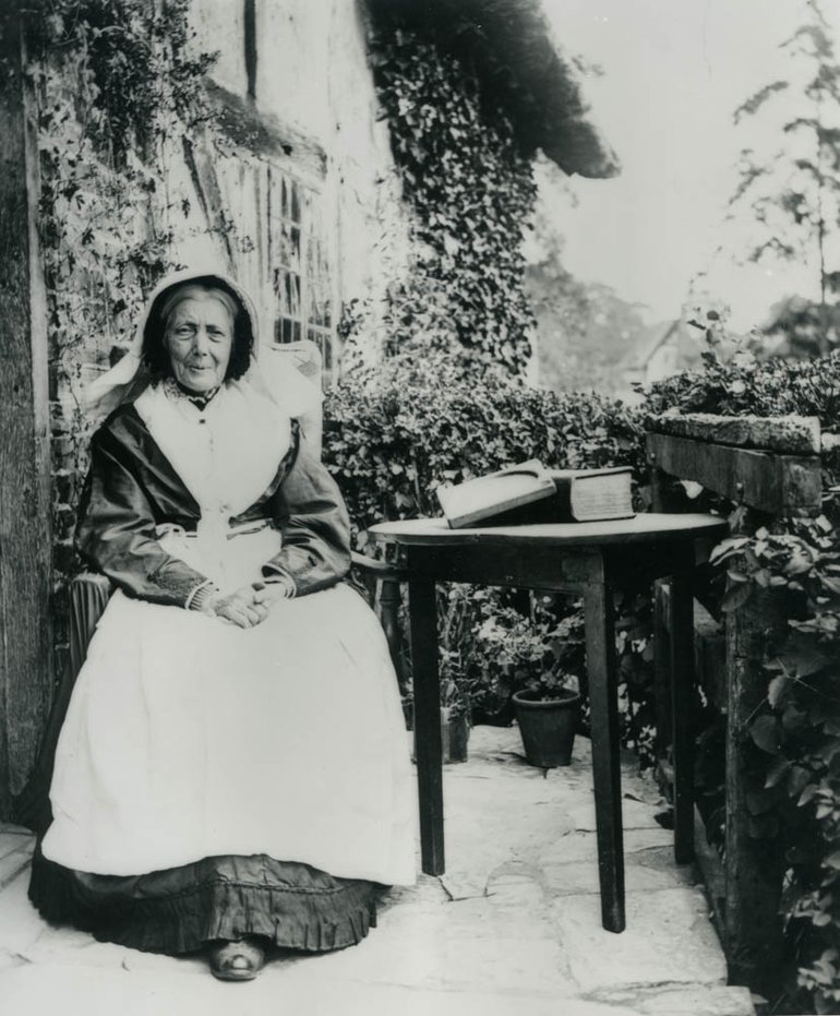 Mary Baker sits outside the cottage door. She wears dark dress, largely covered at the front by a white apron; on her head is a white bonnet, which flows over her shoulders, a bit like a nun's wimple.. On her left is a wooden table with two books, one open resting on the other, which is very fat (probably a Bible). She has an elderly face with half-smile which looks welcoming.