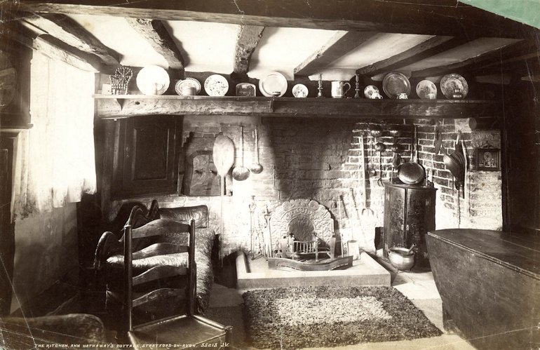 On the raised hearthstone stands a modern metal fire basket in front of a metal fireback; the back wall is soot-stained. On a shelf over the fireplace stand plates and pots. Still in the fireplace but not on the hearth is a corner cupboard on the right, and a seat on the left, half obscured by a chair in front of it. In the foreground on the right is part of a dresser which appears to have a drop-leaf.