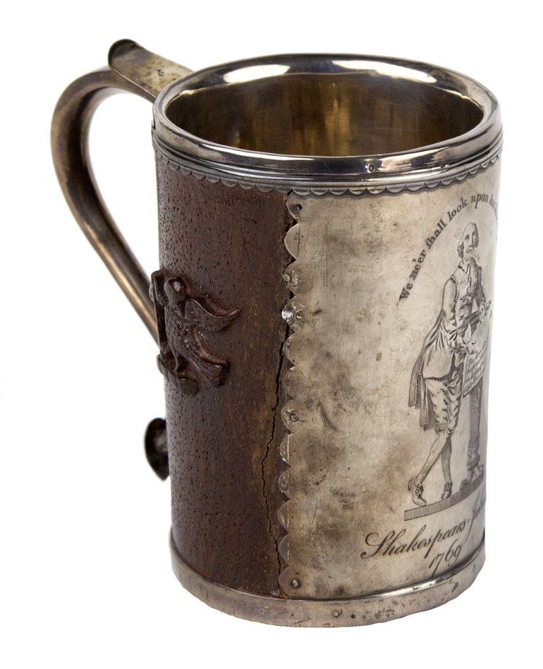 A fairly standard straight-sided tankard with a handle, metal with probably silver trim round the base and top. A full-height plate is on the front, with an engraving of the Jubilee statue (Shakespeare leaning on a pillar, unrolling a manuscript in front of the pillar).