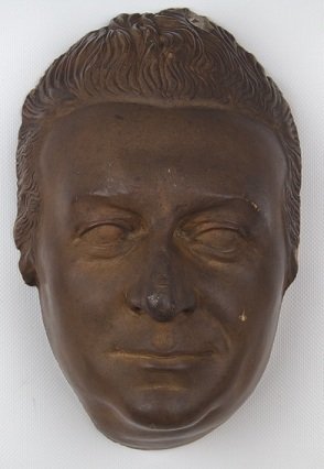 The mask in a dark material, shows him with swept-back curly hair and a high forehead. The face is fairly long, with a rounded but narrow chin.