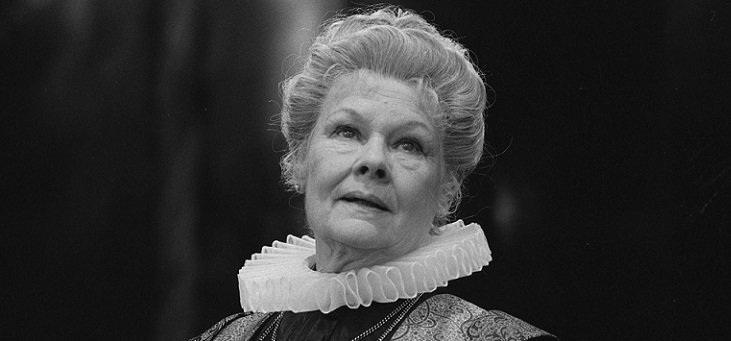 The head of Dame Judy Dench, with curled blond hair and wearing a ruff looking heavenward with a worried expression.