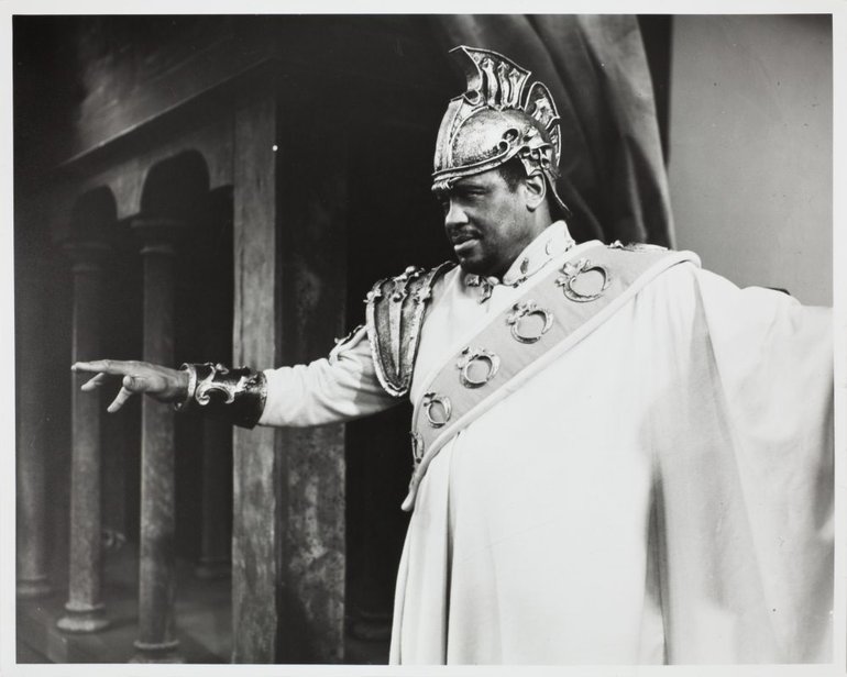 Paul Robeson as Othello, Shakespeare Memorial Theatre, 1959. He wears a military uniform with a sash from his left shoulder to his waist, a large epaulette on his right shoulder, and a helmet with a metal crest.