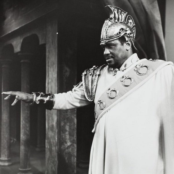 Against the background of a classical arcade, a black Othello, wearing a crested metal helmet, a uniform coat and a sash decorated with a series of rings diagonally over his left shoulder, is pointing to something off to the left (1959 production).