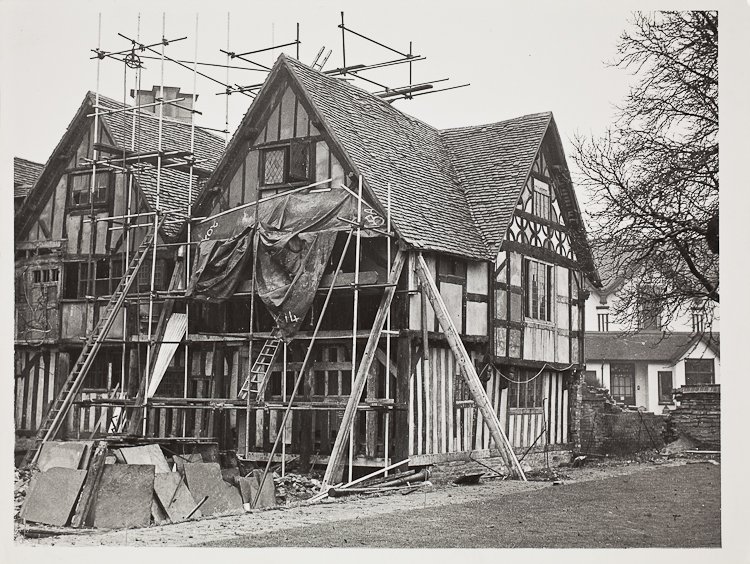 The front and side of Hall's Croft, with scaffolding at the front and across the roof. Much of the front wall has lost its panels and windows, and there is a pile of slabs in the foreground.